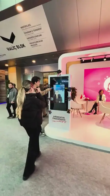 Kongre merkezinde pembe tonlu standda AI photo booth totemine yürüyen ziyaretçi