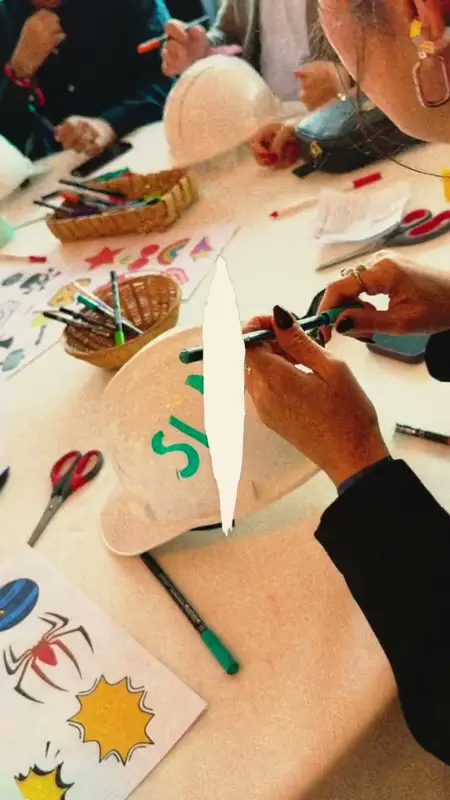Participant writing a message in green letters on a white hard hat at a table