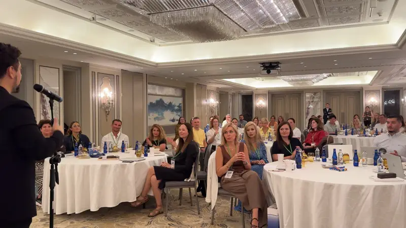 Mikrofonlu kolaylaştırıcının karşısında yuvarlak masalarda oturan kalabalık katılımcı grubu