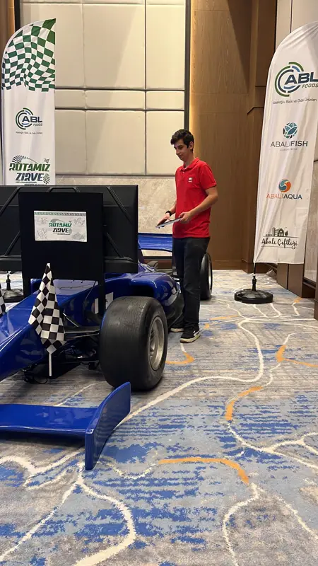 Damalı bayraklar ve kurumsal bayraklar arasında ekranlı mavi F1 simülatörü ve tabletli M3 Works görevlisi