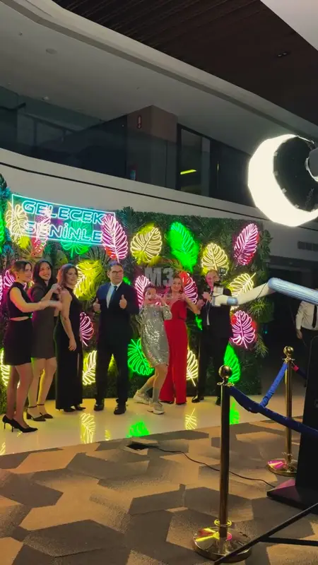 Neon yapraklı duvarın önünde poz veren misafirler, yanda yuvarlak ışık ve kordonlu bariyer
