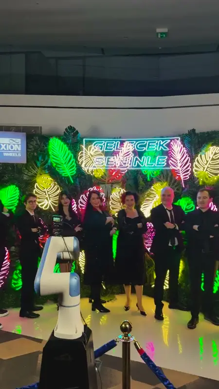 Neon yapraklı fotoğraf duvarının önünde poz veren ekip ve önde kameralı robot kol