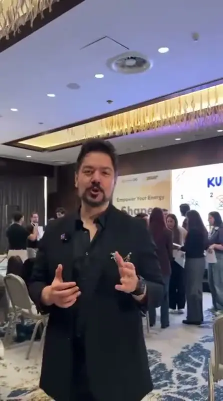 Otel salonunda mikrofonla Human Bingo oyununu anlatan sunucu, arkada bingo kâğıtlarıyla dolaşan katılımcılar