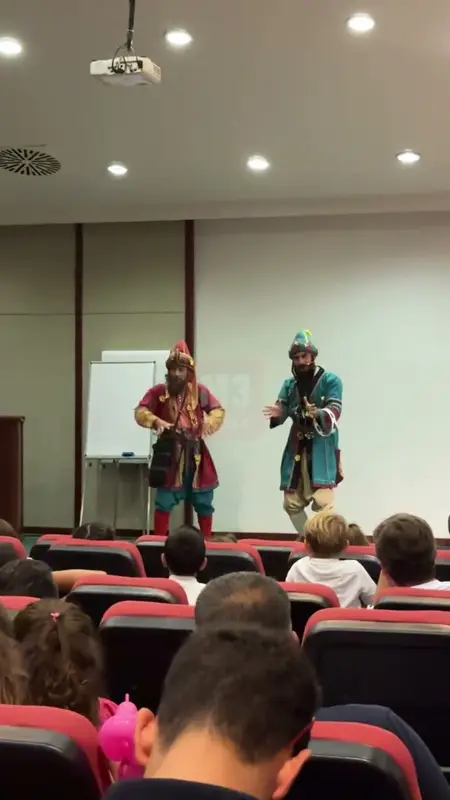 Two actors in Karagöz and Hacivat costumes on a conference hall stage with children watching
