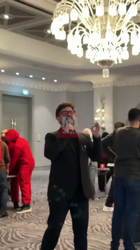 Otel salonunda mikrofonla oyunu yöneten sunucu ve arkada kırmızı tulumlu katılımcılar