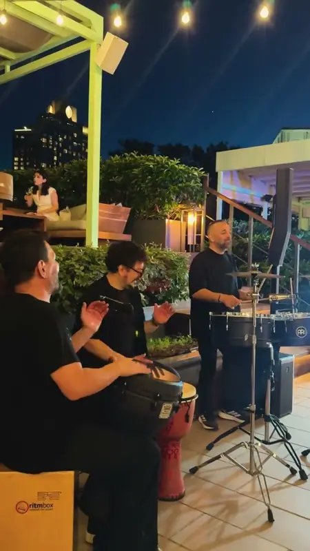 Percussionists playing hand drums under evening lights, with timbales behind them