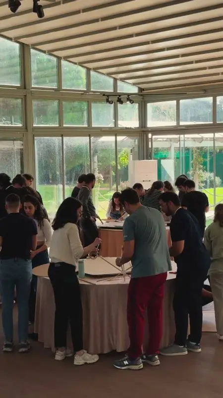 Cam duvarlı salonda yuvarlak masalar etrafında robot inşa eden takımlar