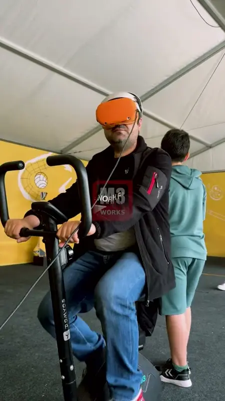Sabit bisiklette VR gözlüğü takmış yetişkin katılımcı, arkasında sırasını bekleyen genç